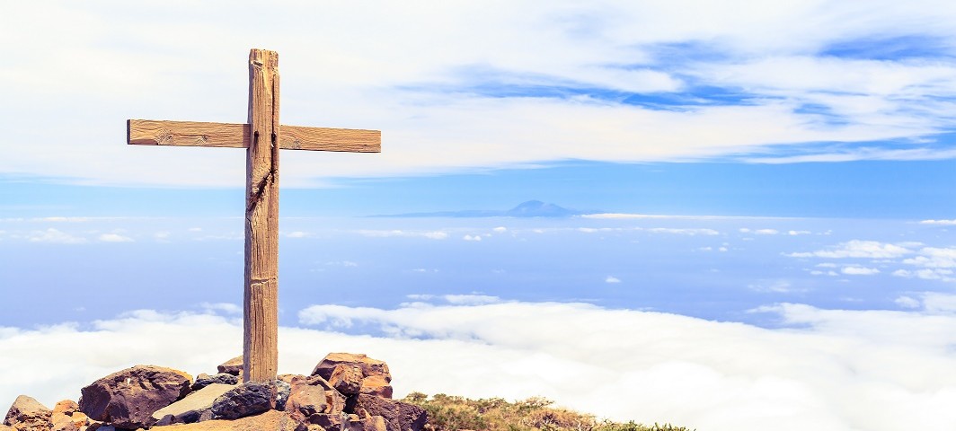 Christian wooden cross on mountain top, rocky summit, beautiful inspirational landscape with ocean, island, clouds and blue sky, looking at scenic blue sea and white clouds.